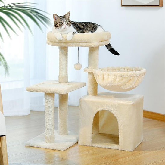chat tigre et blanc sur le couchage supérieur d’un petit arbre à chat beige avec niche et panier suspendu.